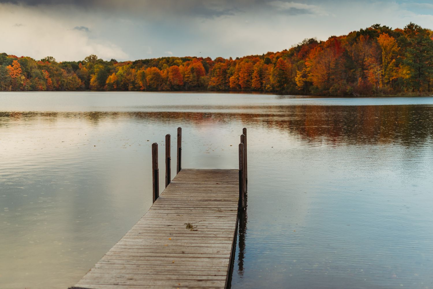 Wooden dock extends into a calm lake surrounded by vibrant autumn foliage at sunset.