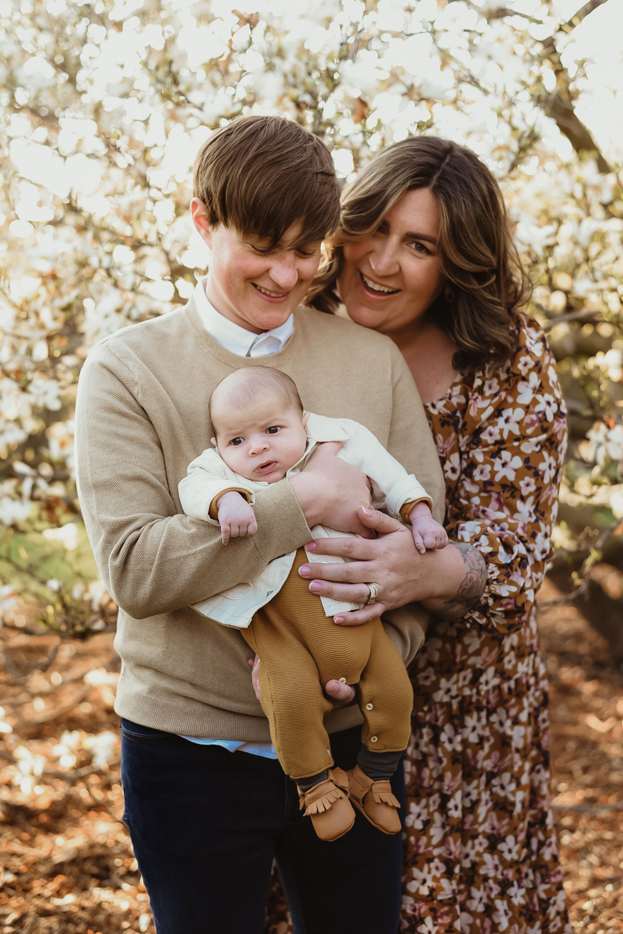 A family poses together in front of blooming spring trees while holding a baby in casual attire.