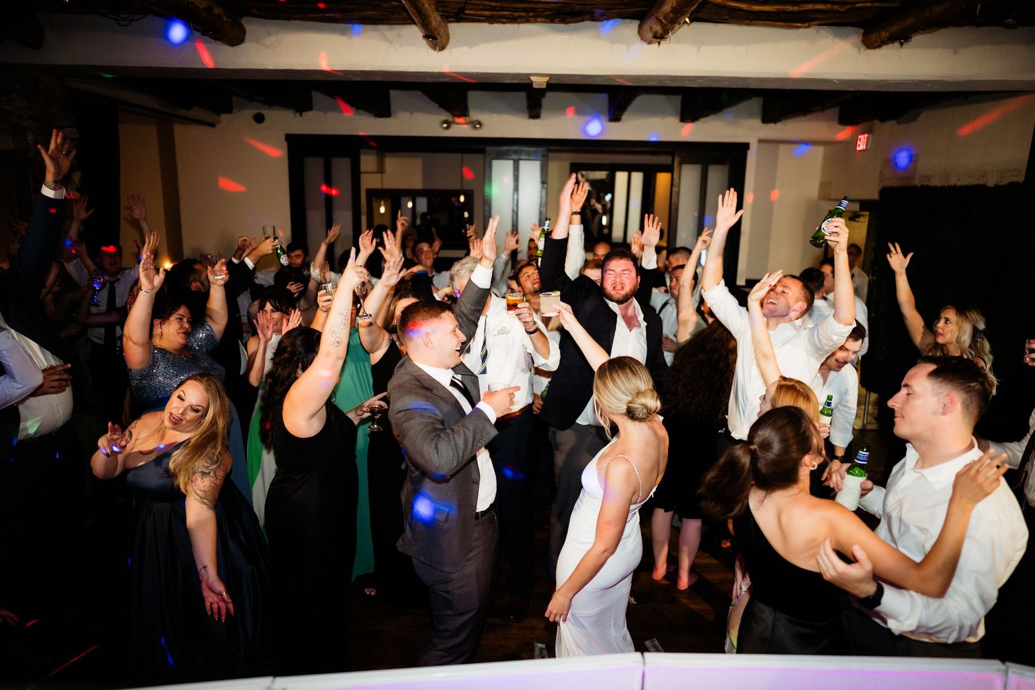 Wedding guests dancing with raised arms on a crowded dance floor with blue lighting.