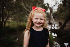 A child with curly hair and red bow smiles brightly near a pond with trees in background.