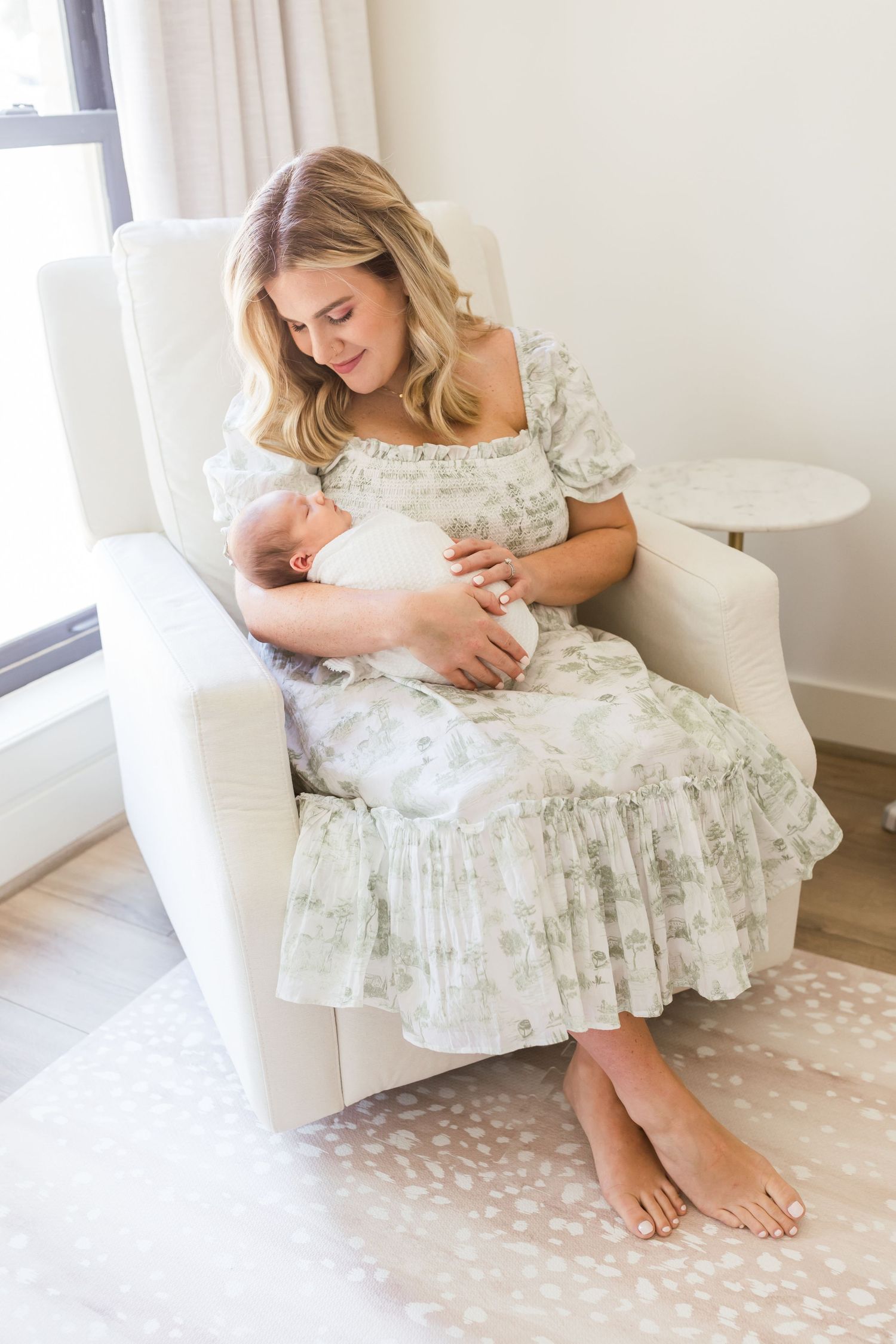 Mother in floral dress cradles newborn while sitting barefoot in white armchair.
