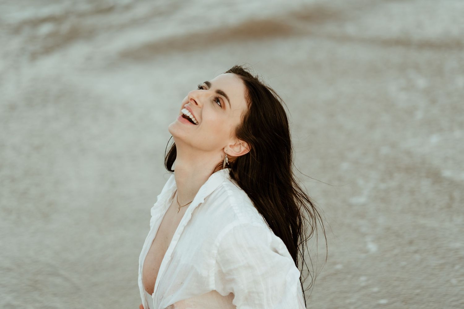 A candid outdoor portrait of someone in white clothing laughing joyfully with windswept dark hair.