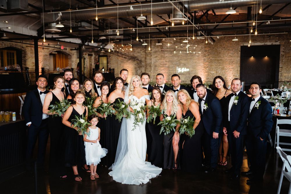 Wedding party members gather for a group photo in an industrial-style venue with exposed brick and hanging lights.