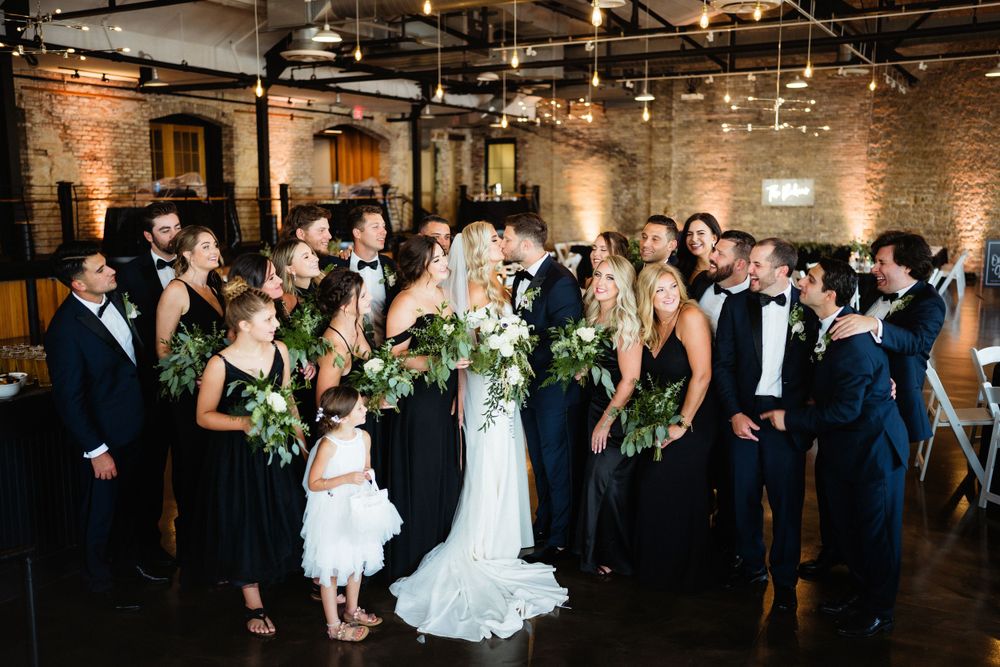 Large wedding party poses together in formal attire against exposed brick wall with string lights.
