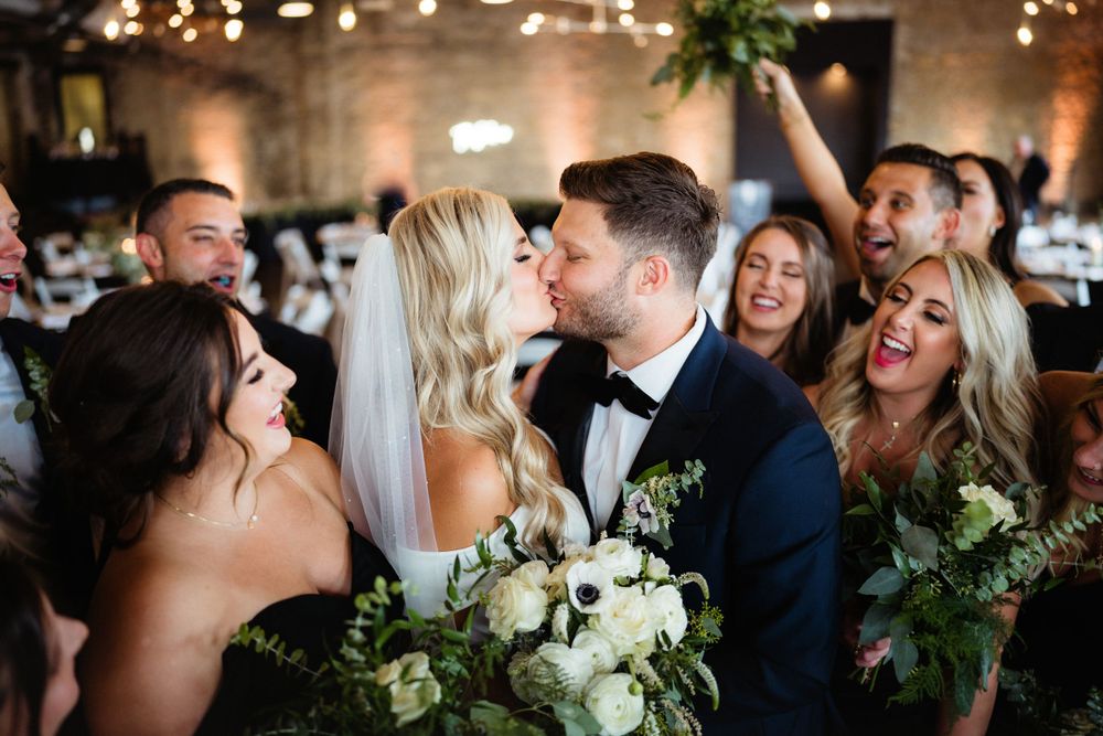 Joyful wedding party celebrates with laughter and white floral bouquets in an industrial venue with brick walls.