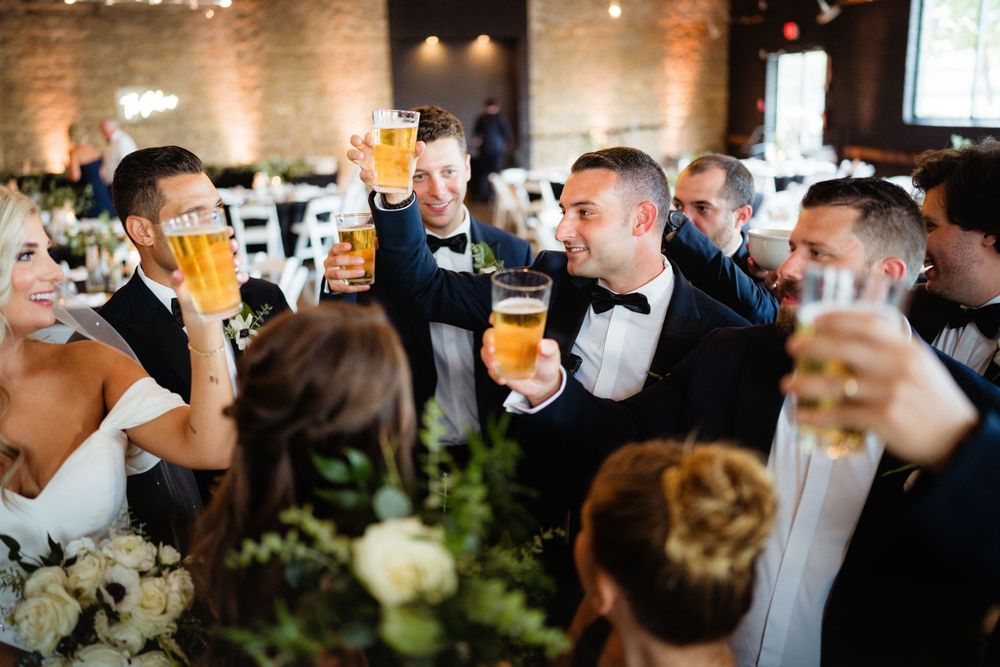Wedding party members clink glasses of beer together in celebration at a rustic reception venue.