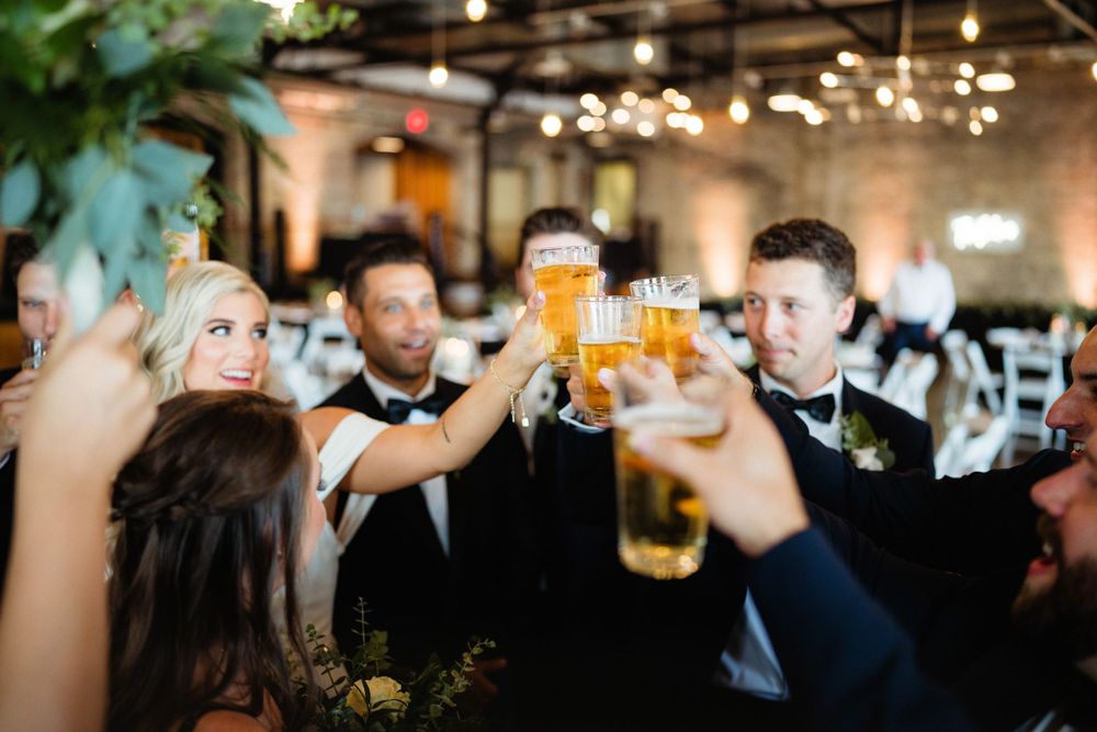 Wedding guests raise beer glasses in a cheerful toast at an industrial-chic venue with exposed brick walls and string lights.