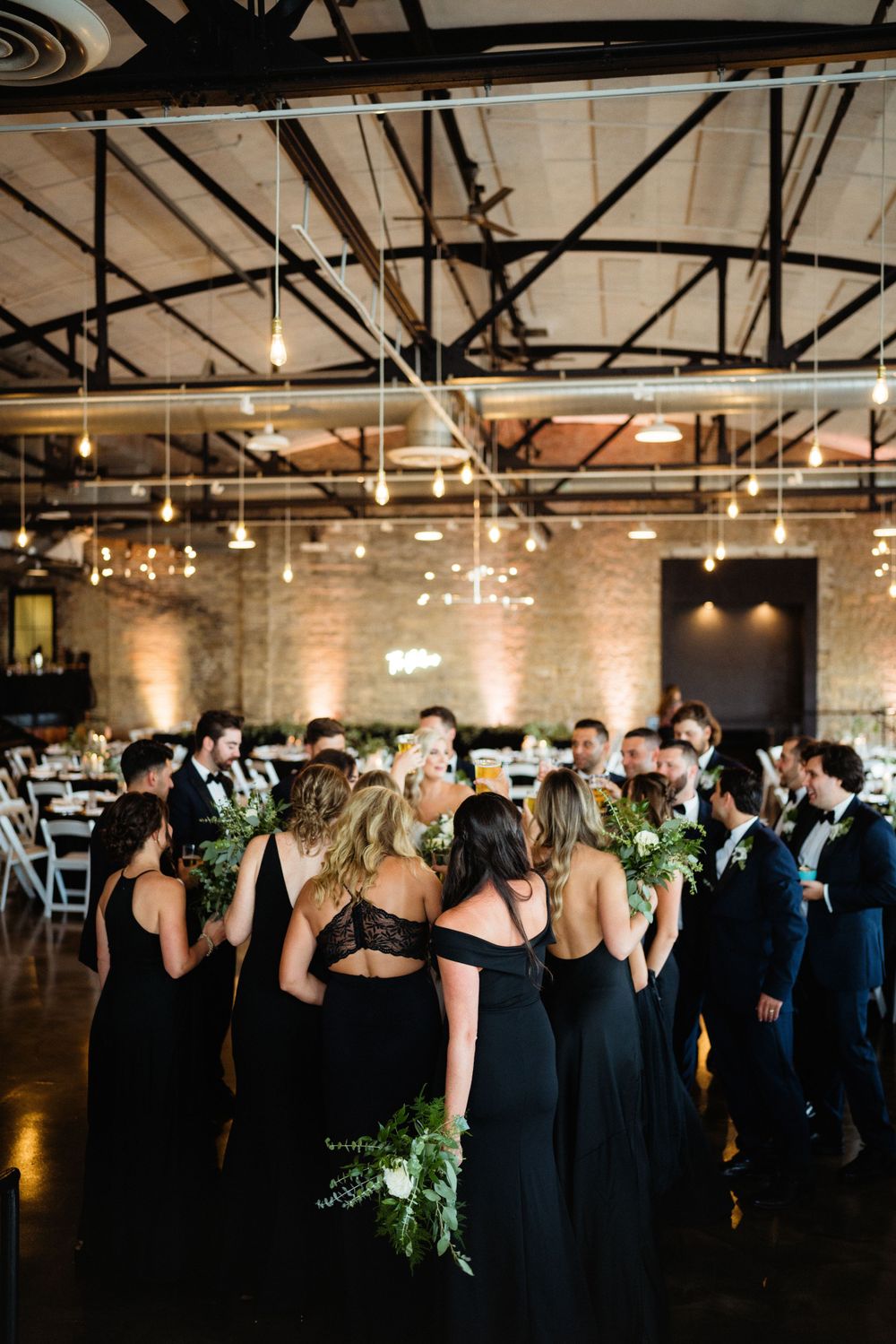 Wedding party in black attire gathers under industrial ceiling with string lights.