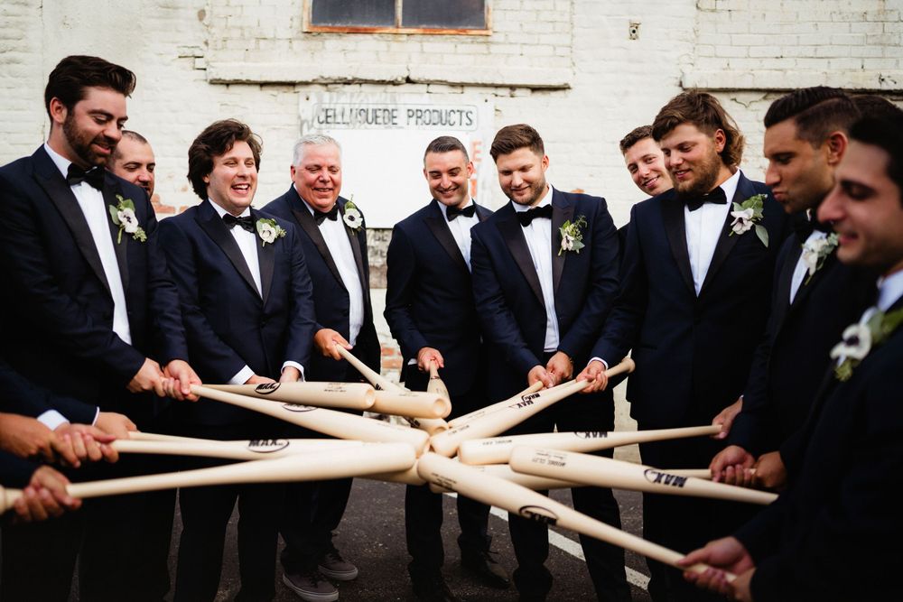 Groomsmen gather in circle holding drumsticks in ceremonial pose outside brick building.