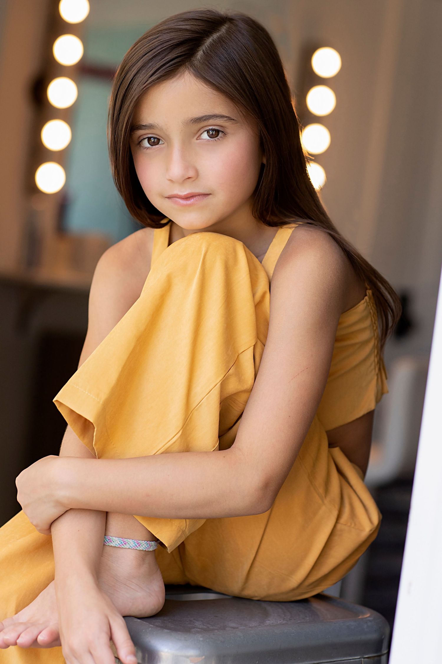 Young performer in yellow dress sitting casually against blurred vanity mirror background.
