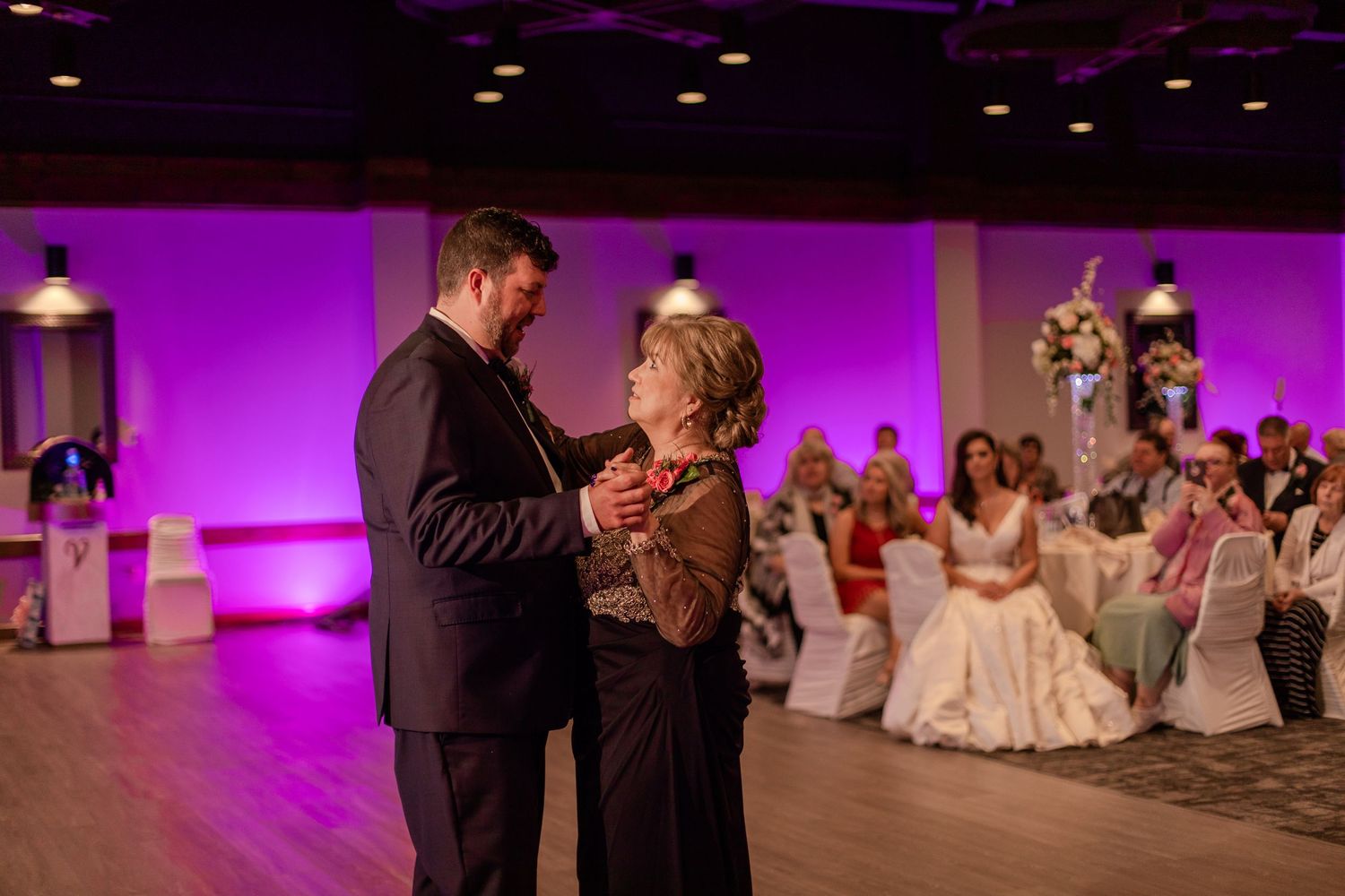 A dance moment captured at a wedding reception with purple ambient lighting and decorated guest tables.