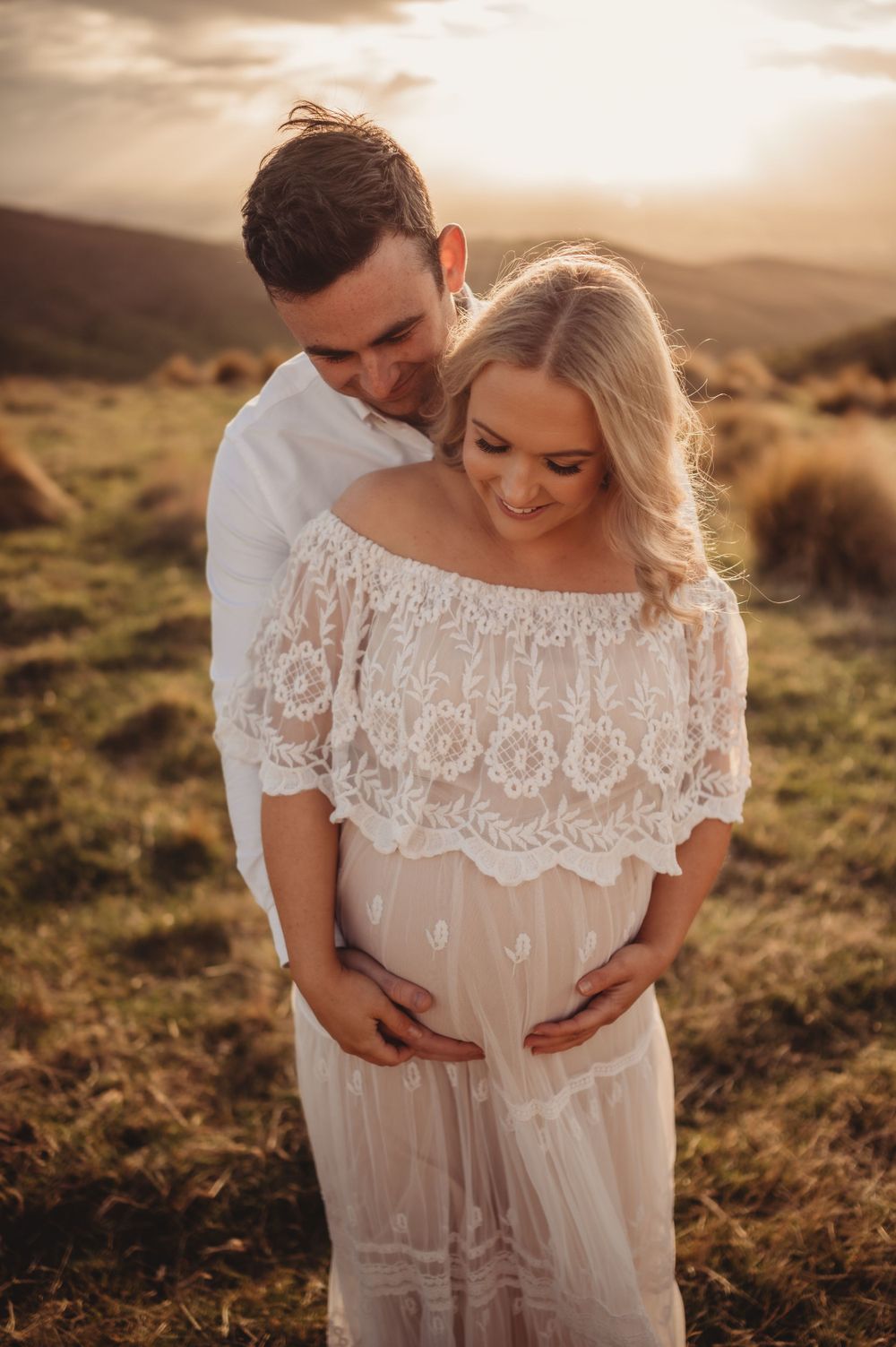 Intimate maternity portrait showing couple embracing in white dress against mountain backdrop.