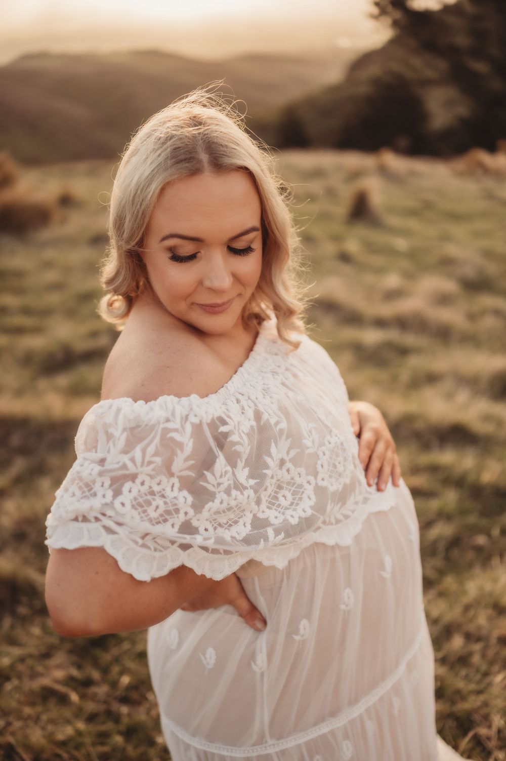 Ethereal maternity photoshoot in white lace dress against golden sunset in mountain landscape.