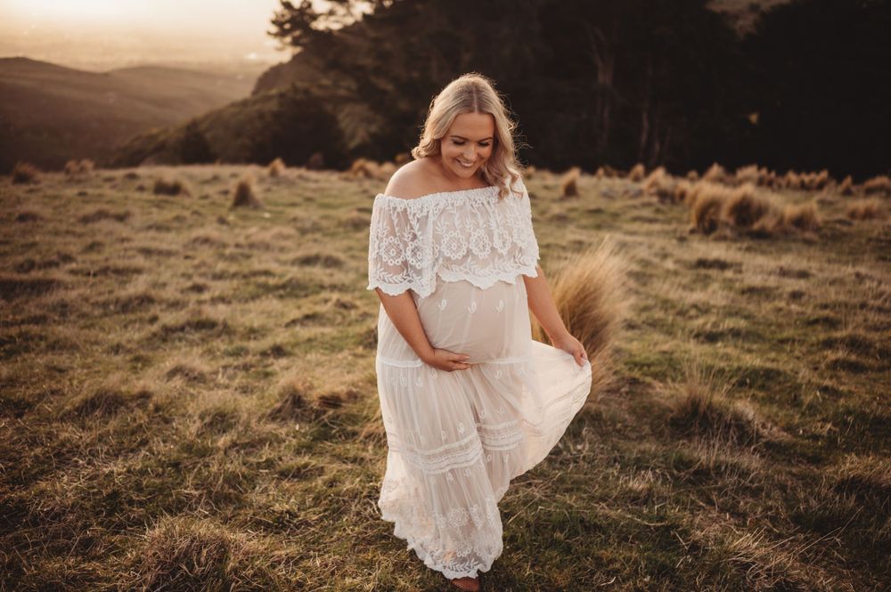 Expectant mother in white lace dress standing in sunlit meadow at sunset.