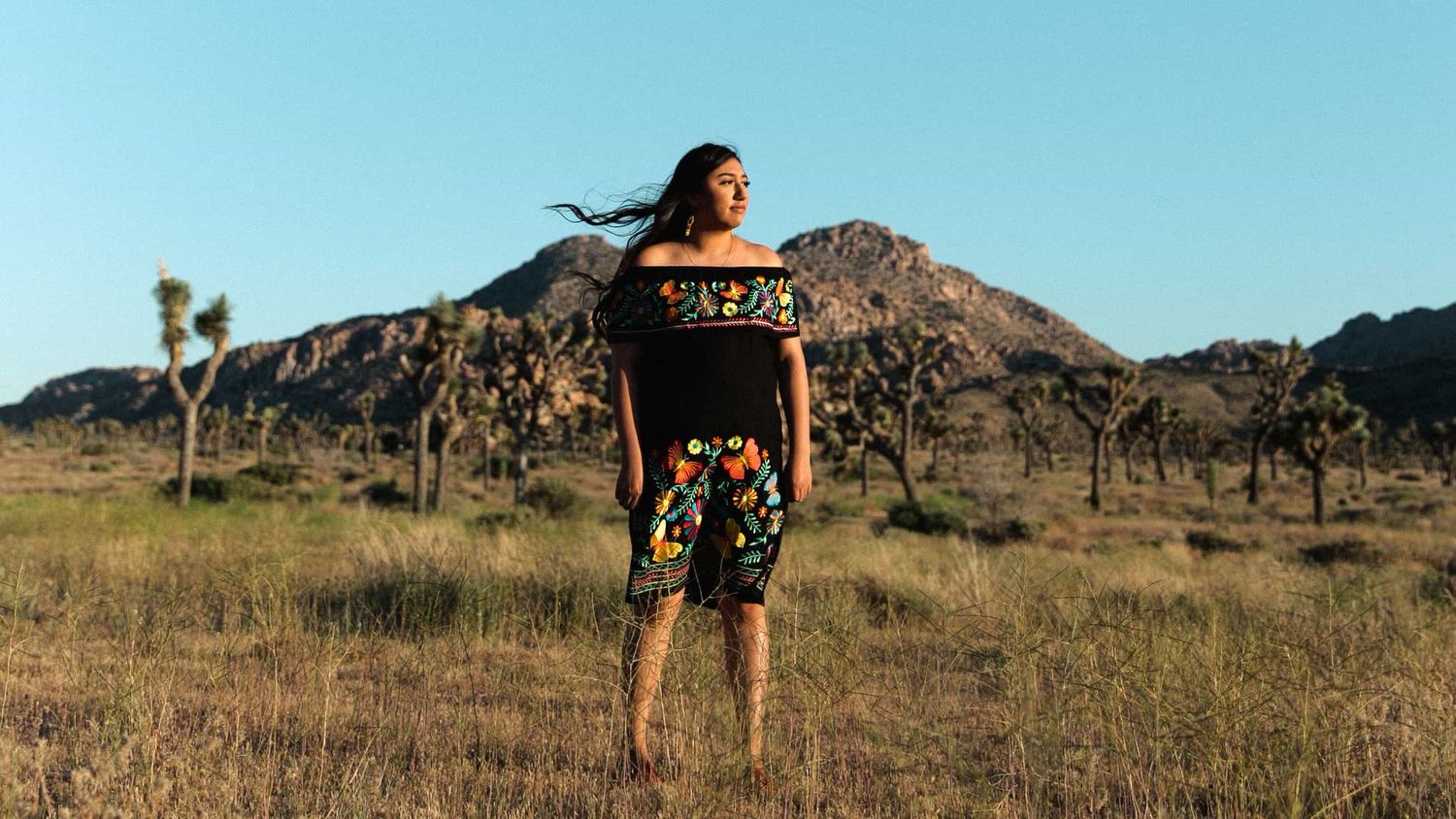 Woman in black floral outfit stands confidently in desert landscape with Joshua trees and rocky hills.