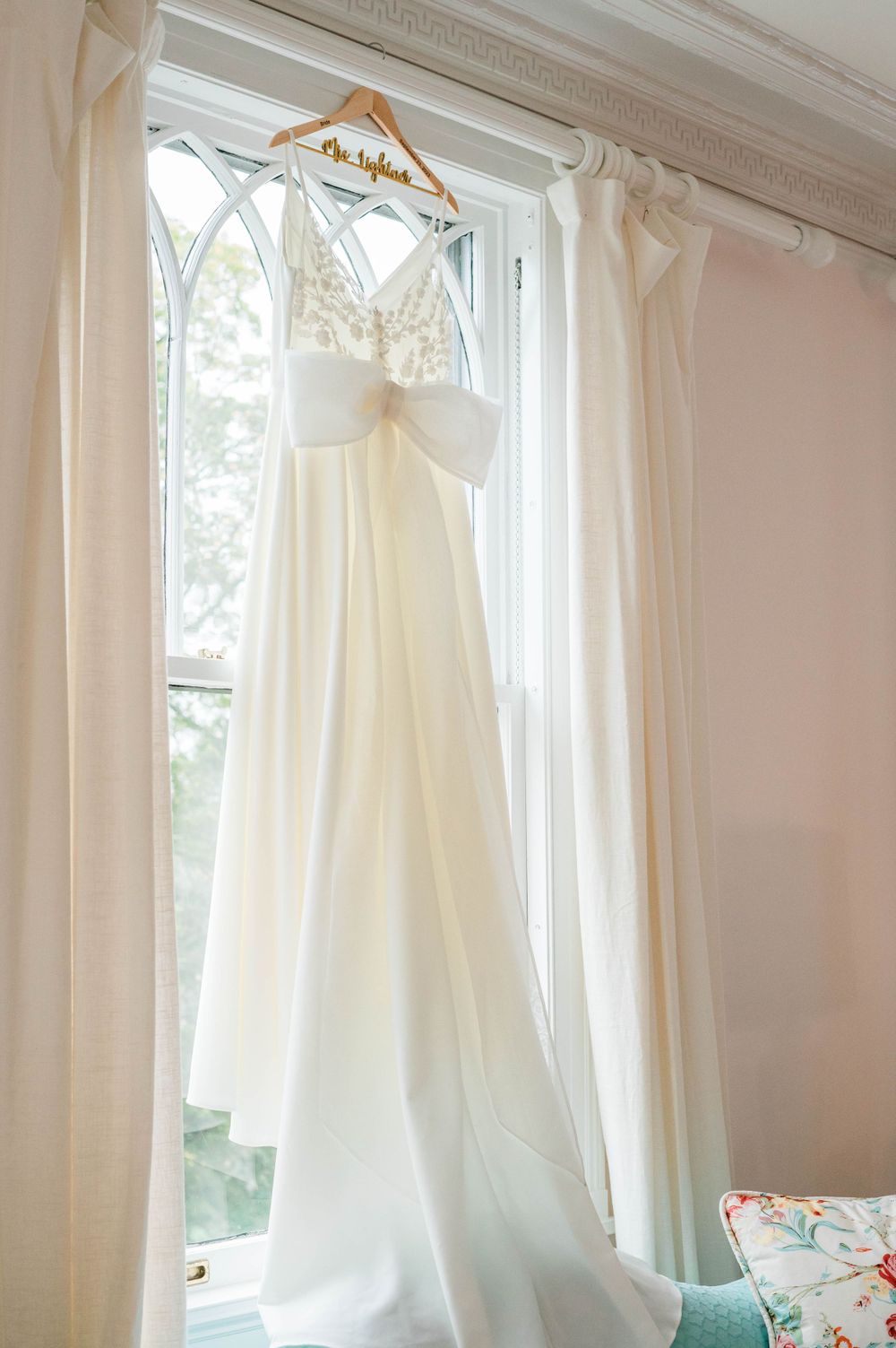 A white wedding dress hangs elegantly in front of a bright window with flowing curtains.