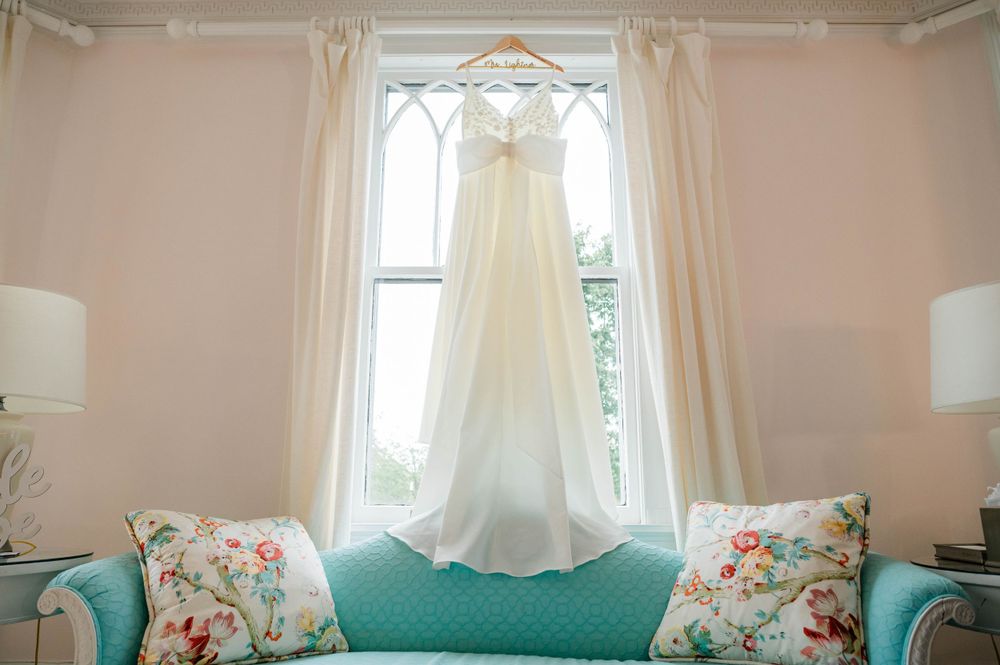 White wedding dress hanging in window with floral accent pillows on turquoise bench below.