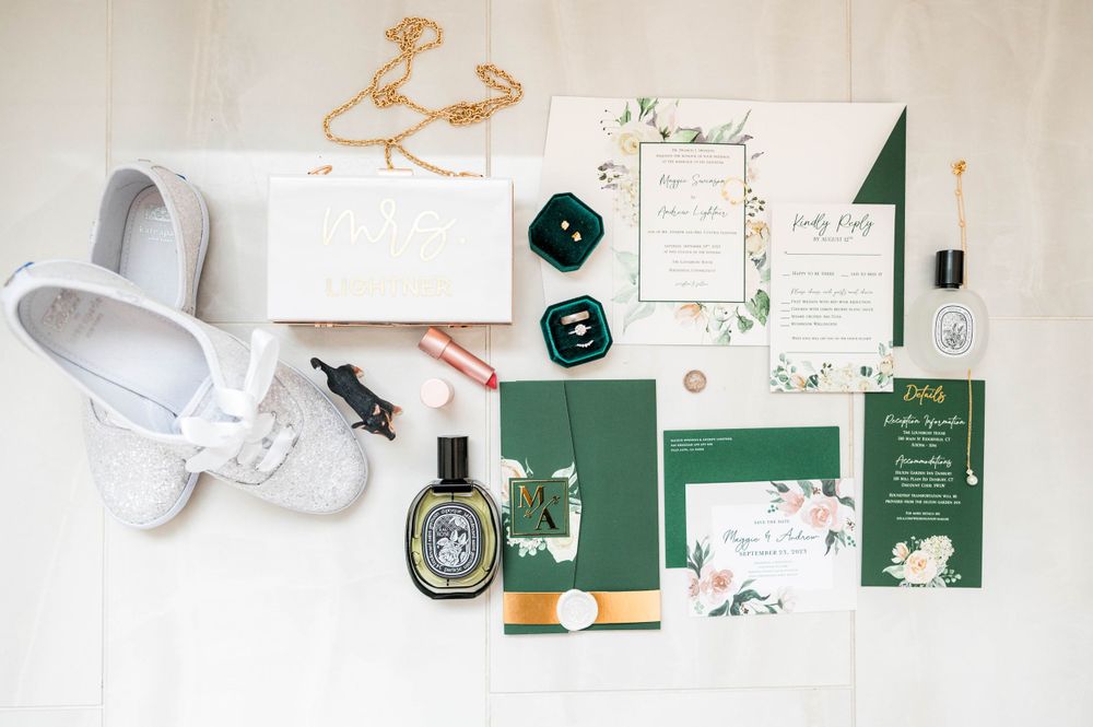 Wedding invitation suite with green accents, white shoes, and perfume bottle laid flat.