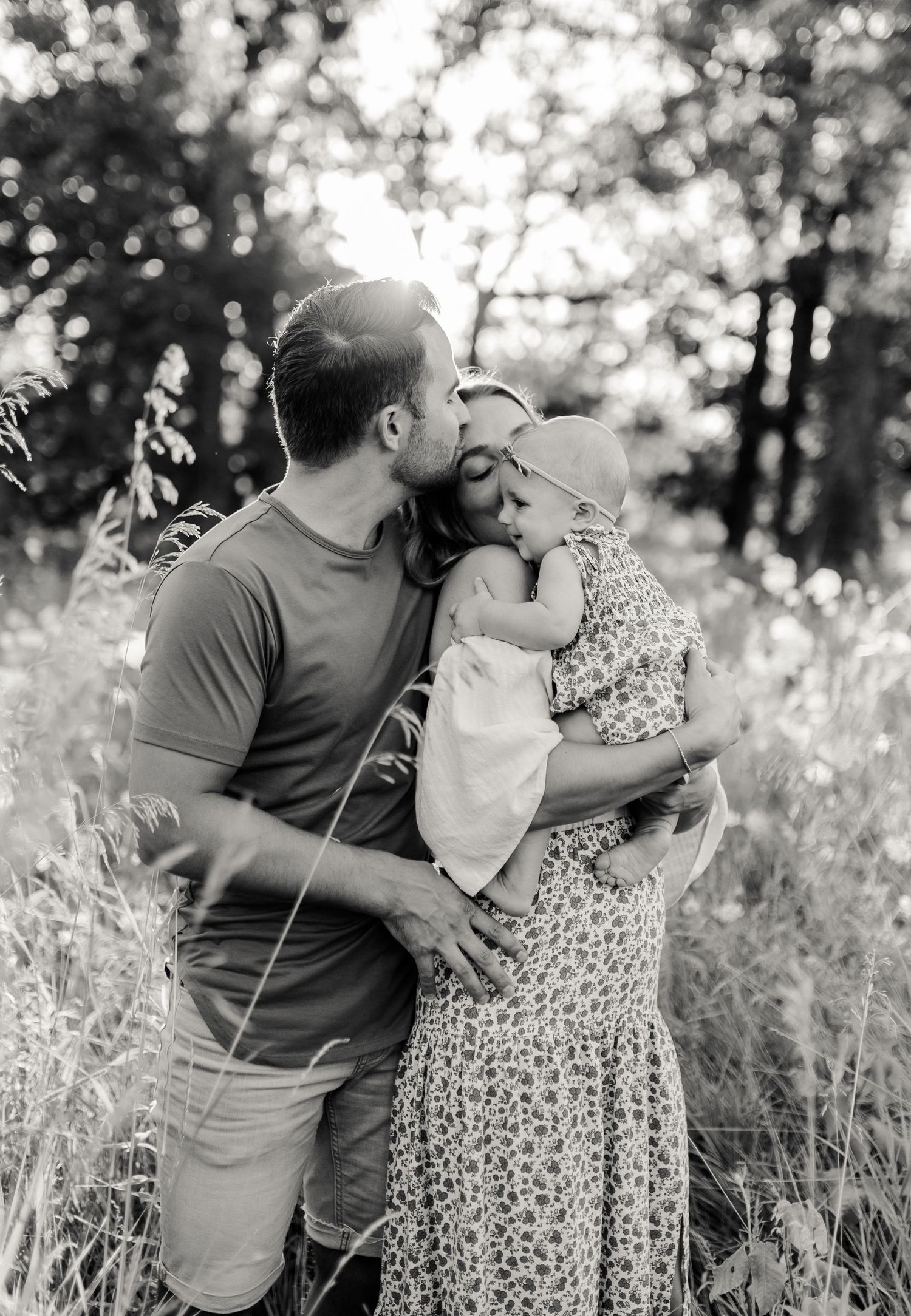 Black and white photo of family embracing in sunlit field with tall grass.