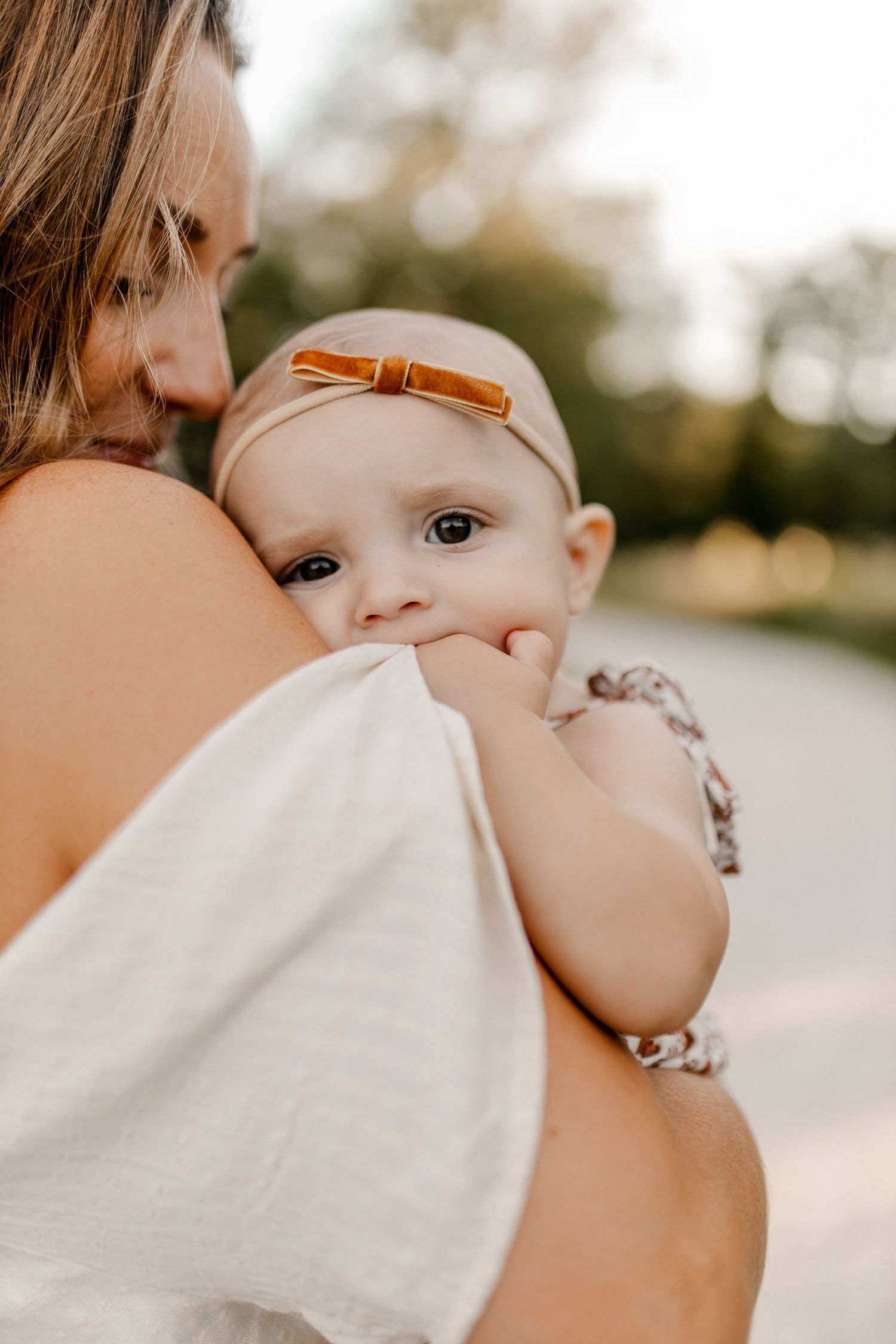 Baby wearing bow headband rests on parent's shoulder in warm sunlight.