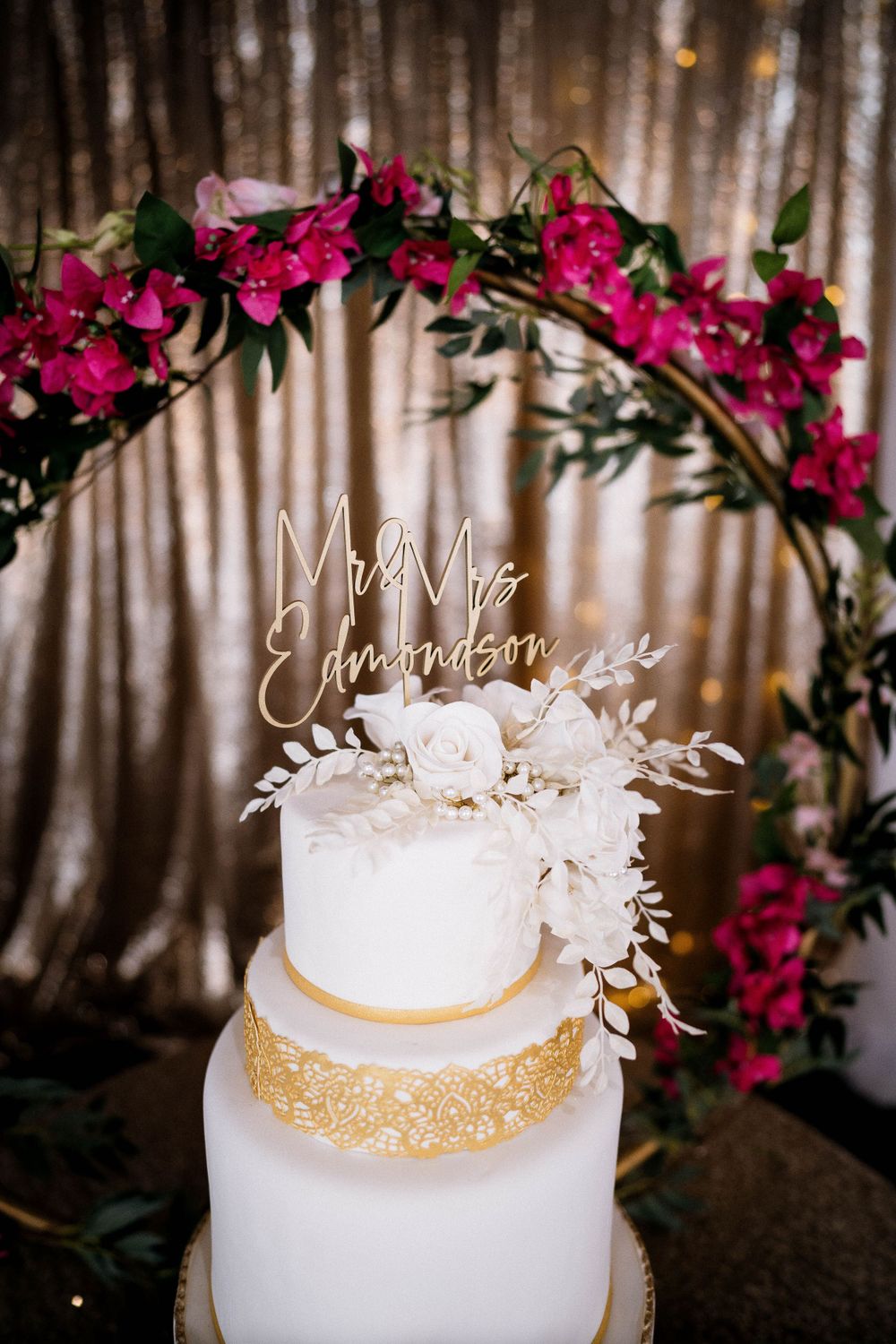 Elegant three-tier white wedding cake with gold accent and pink floral arch topper.