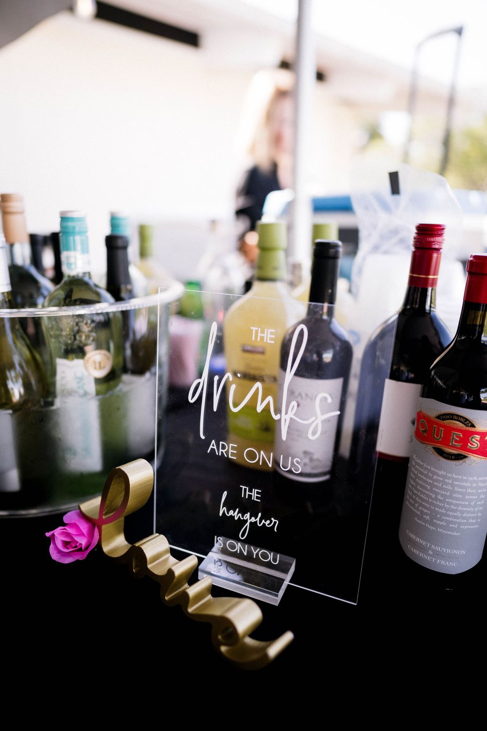 Bar setup with wine bottles and acrylic sign reading 'The drinks are on us' at wedding reception.