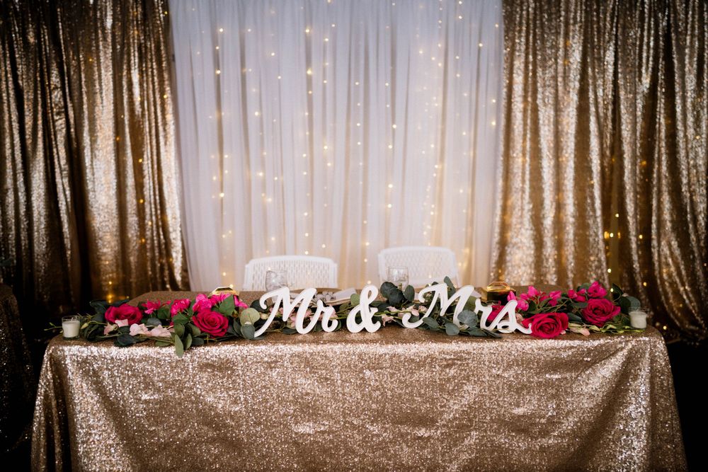 Wedding reception backdrop with gold sequin tablecloth and Mr & Mrs sign decorated with pink flowers.