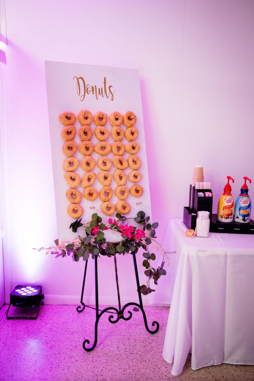 A wedding donut wall display with pink lighting and floral arrangement features rows of glazed donuts.