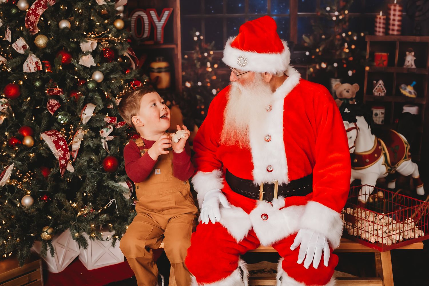 Young child sits with Santa Claus next to decorated Christmas tree with lights and ornaments.