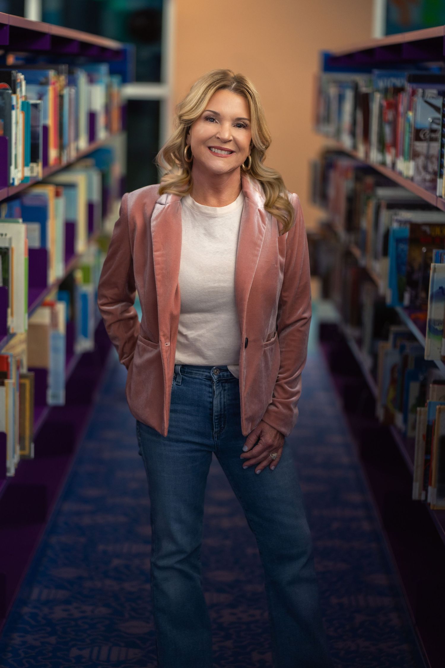 Smiling blonde haired female author in pink blazer surrounded by children's books in a library