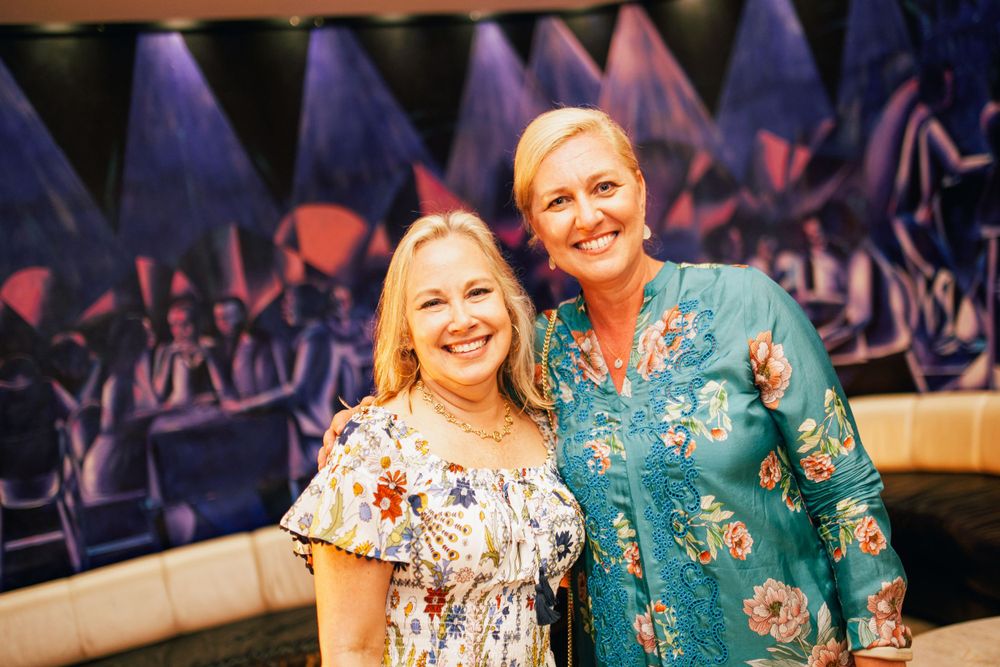 Two people wearing floral dresses pose together at an evening event.