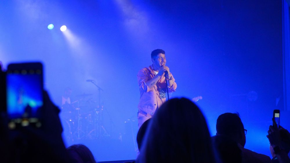 Performer on stage illuminated by blue spotlights while audience members record on phones.