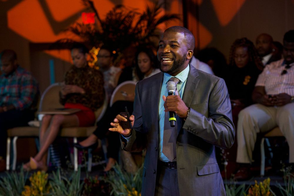 Speaker in suit delivers presentation in warm-lit venue with audience visible in background.