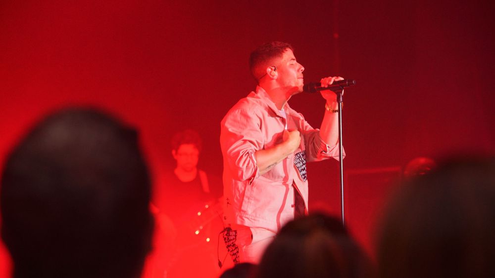 Stage performer in light colored jacket singing under dramatic red stage lighting.
