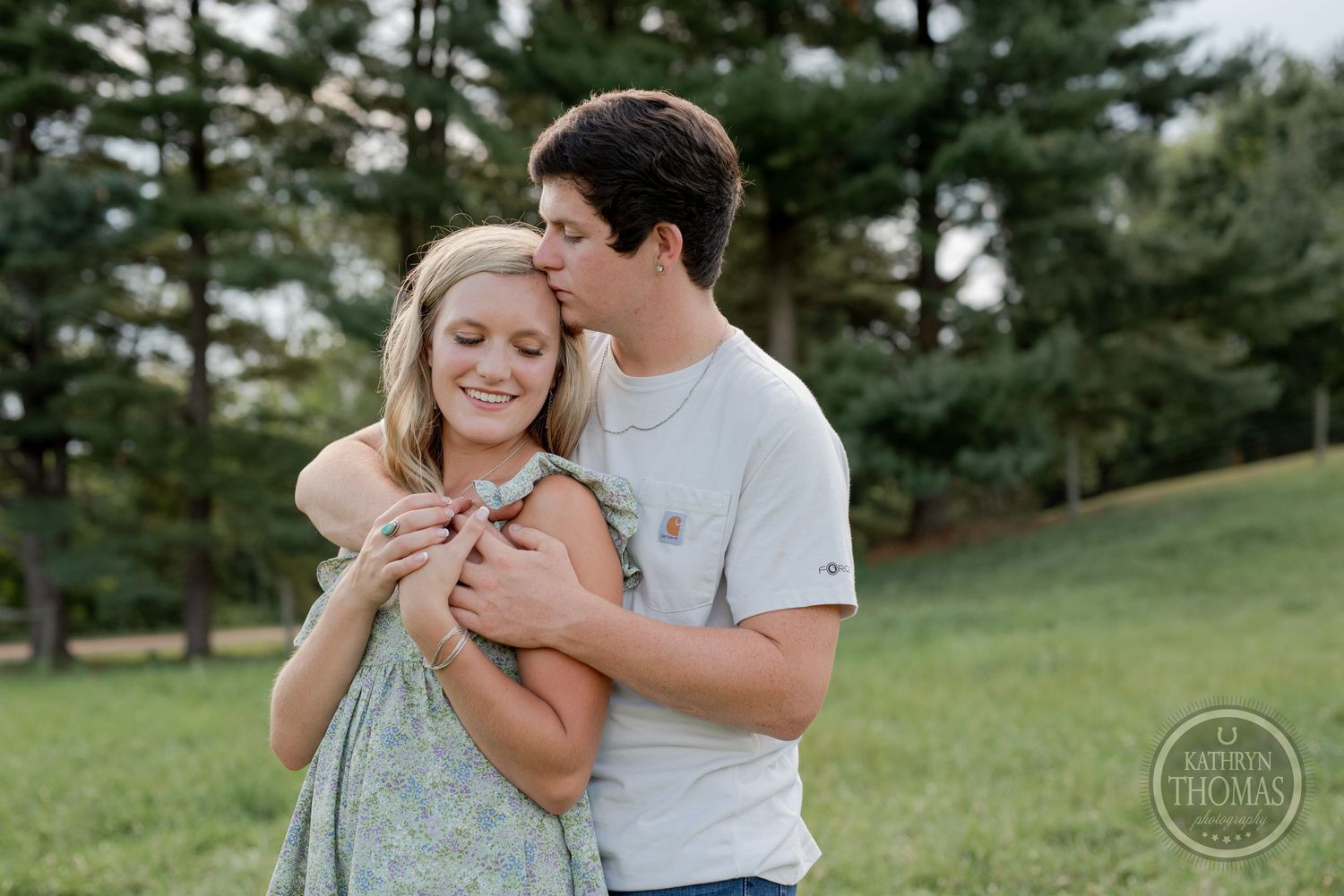 Romantic couple embraces in grassy field with pine trees in background.