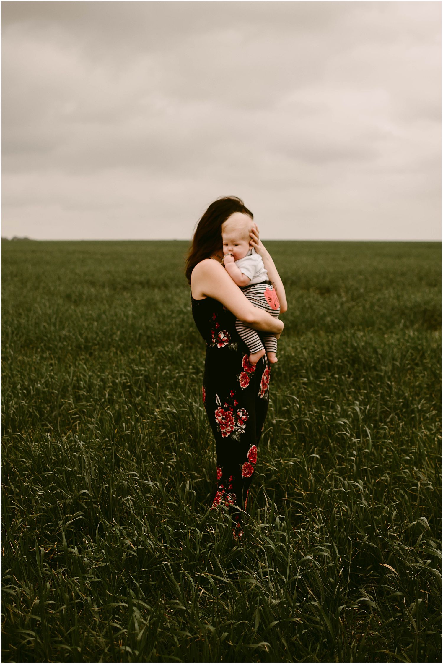 A woman in a floral dress holds a baby while standing in a vast green field.