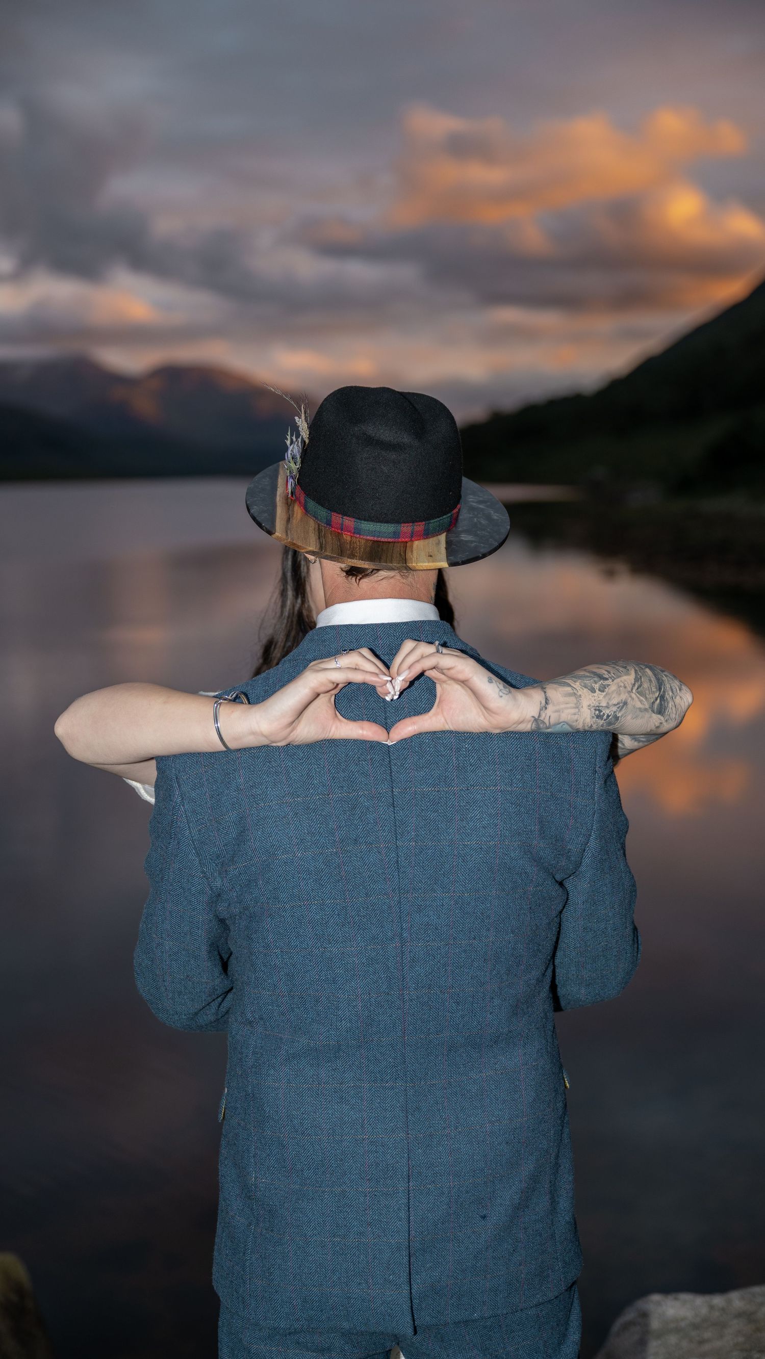 Person in blue suit makes heart shape with hands against sunset lake view.