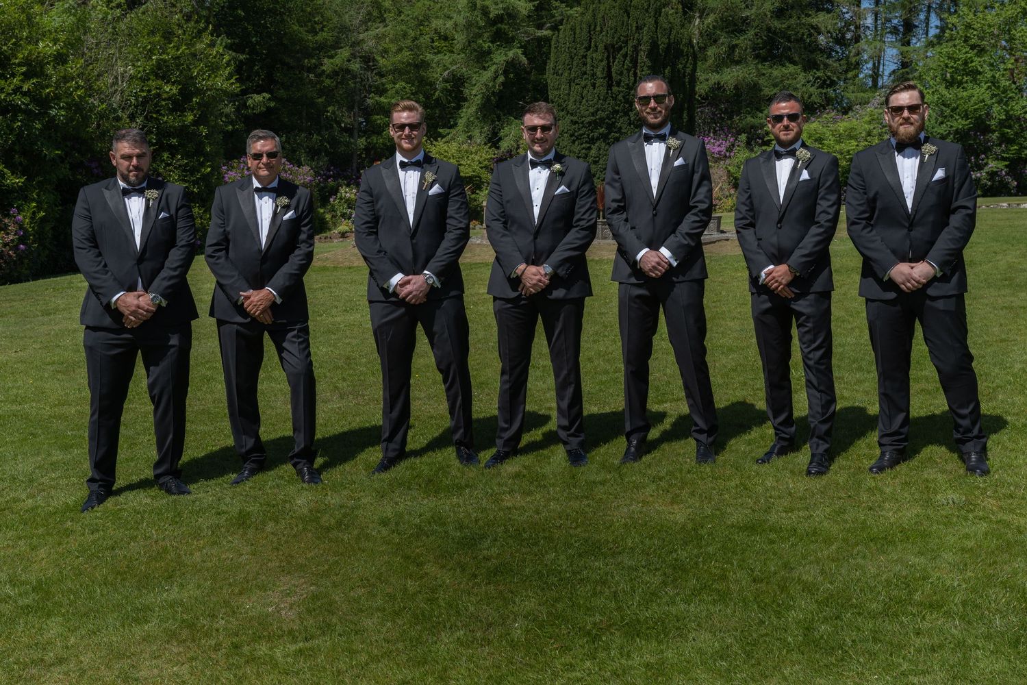 Groomsmen in black tuxedos line up on green lawn for formal wedding photo.