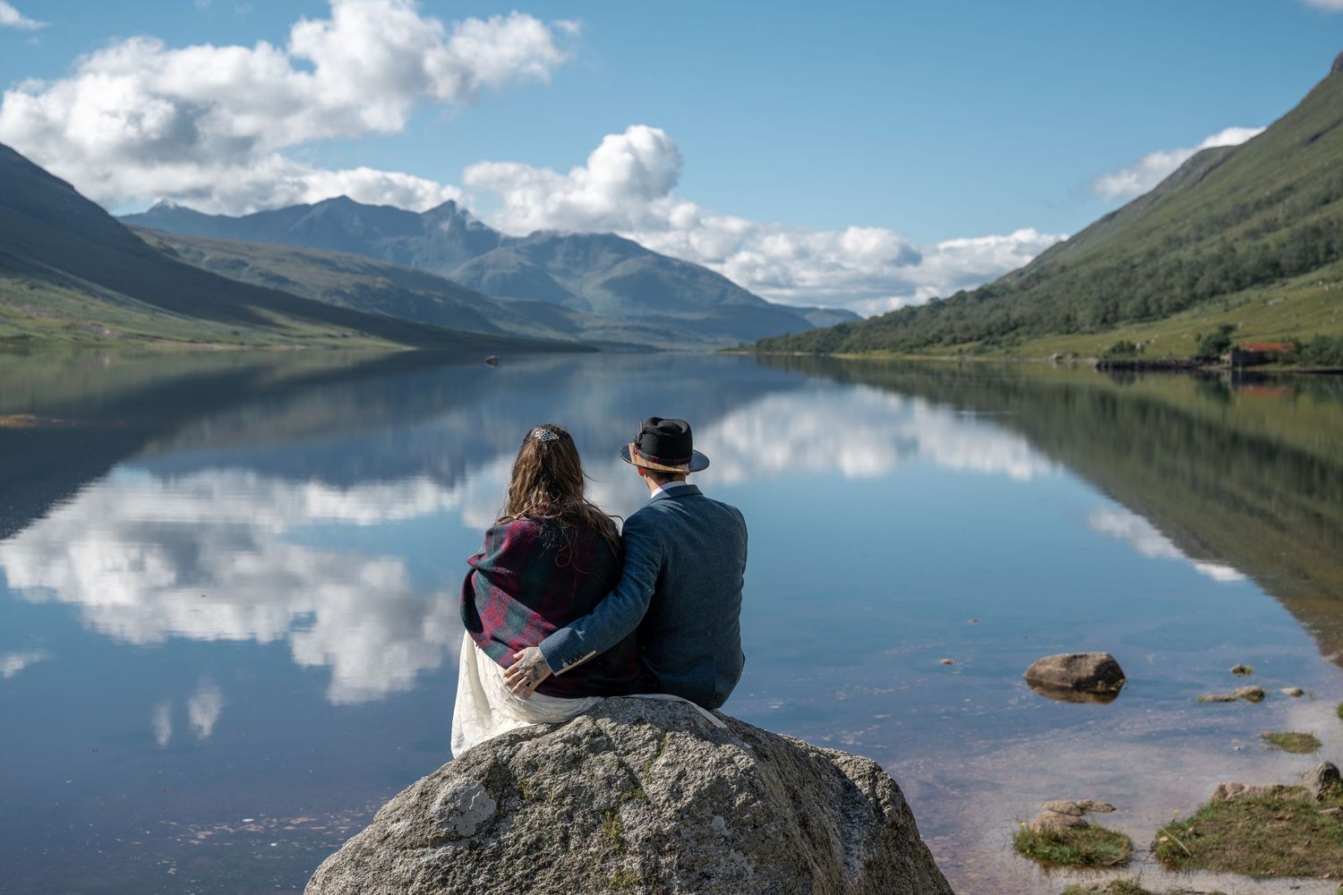 Couple sits together on rock overlooking serene mountain lake on sunny day.