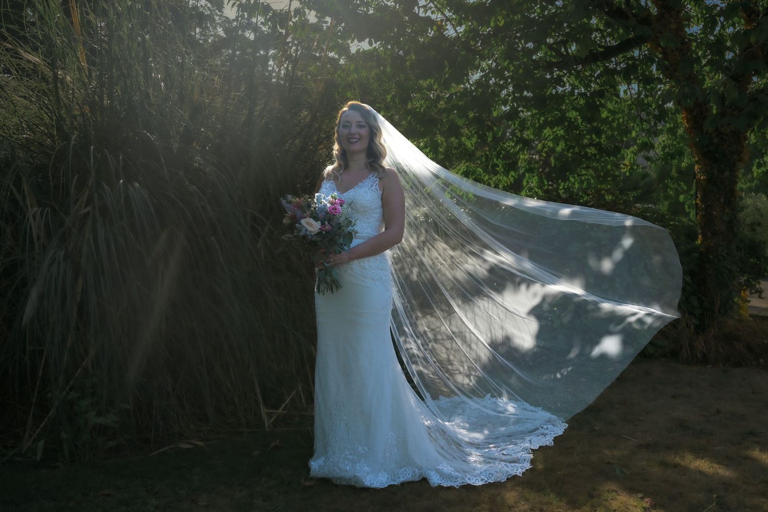 Elegant lace wedding dress and flowing veil catch the sunlight in a romantic forest setting.