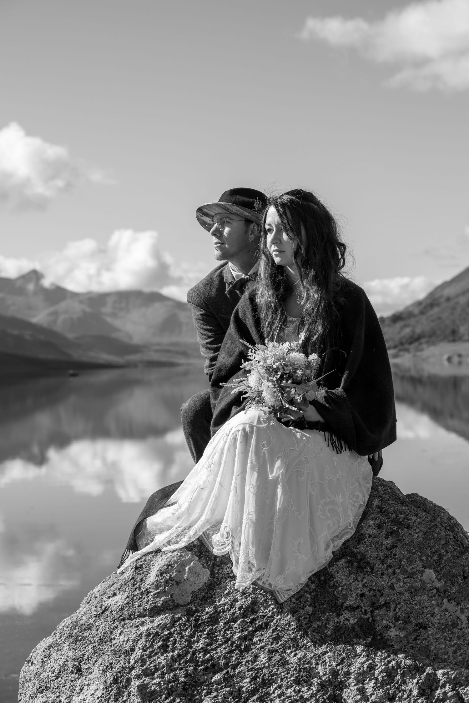 A couple sitting on a large rock by a tranquil mountain lake.