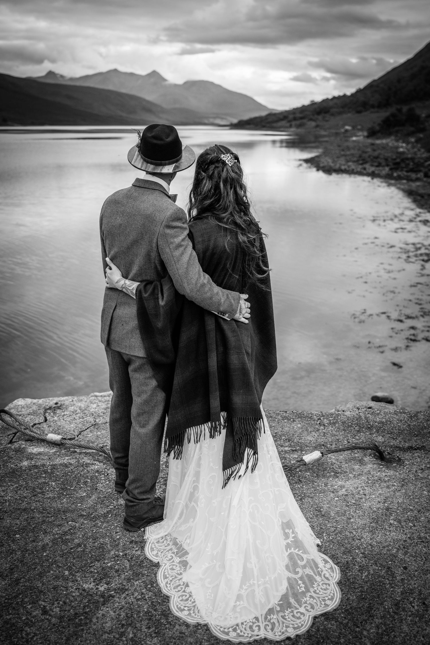 A black and white photo of a couple standing by a serene mountain lake.