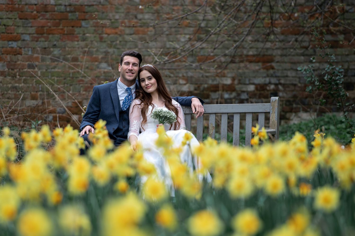 Couple sitting on wooden bench surrounded by blooming yellow daffodils against brick wall.