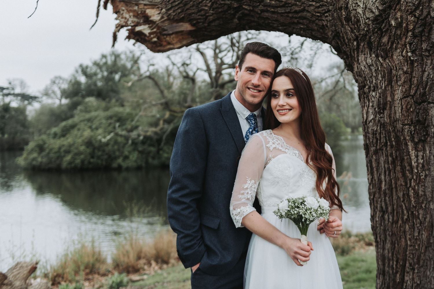 Wedding couple standing close together under a large tree branch near a lake.