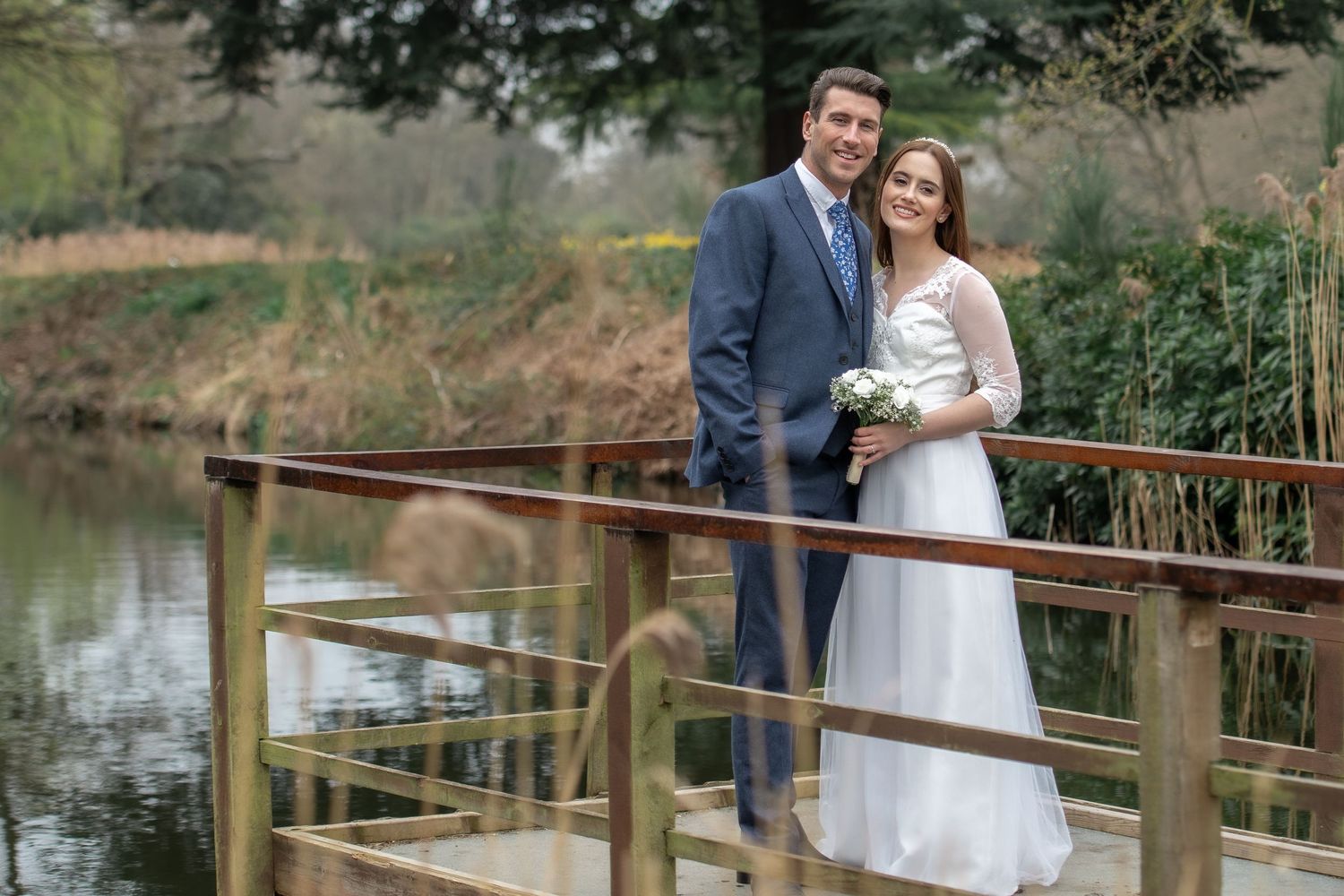 Wedding couple standing on wooden bridge overlooking pond in natural setting.