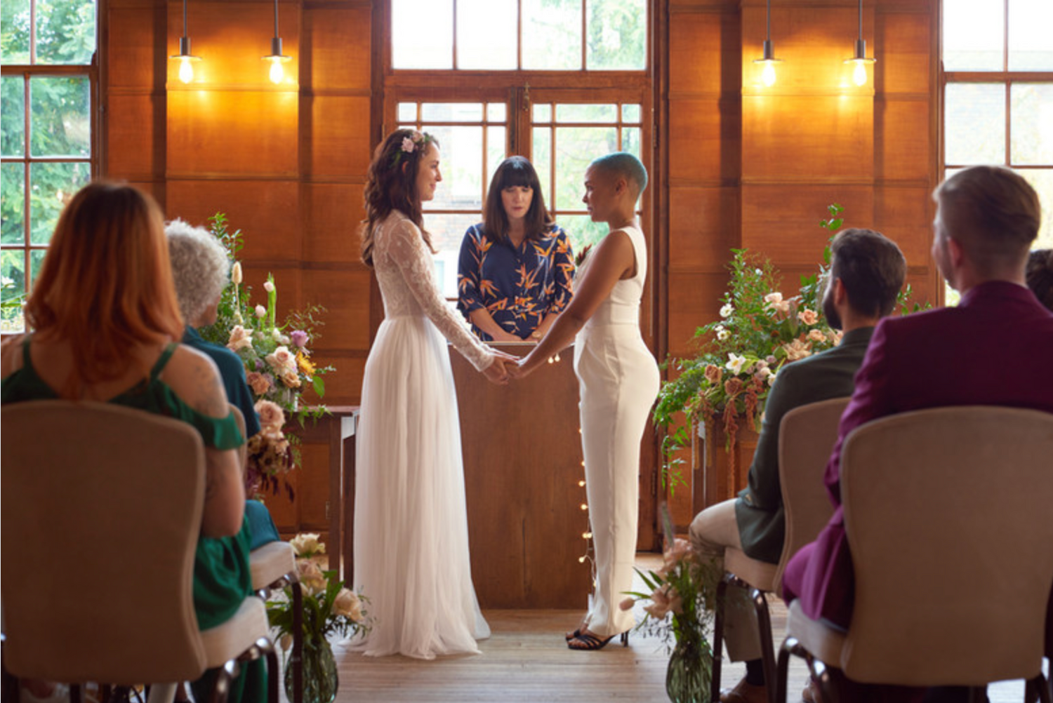 Indoor wedding ceremony in a warm wooden room with floral arrangements and guests seated.