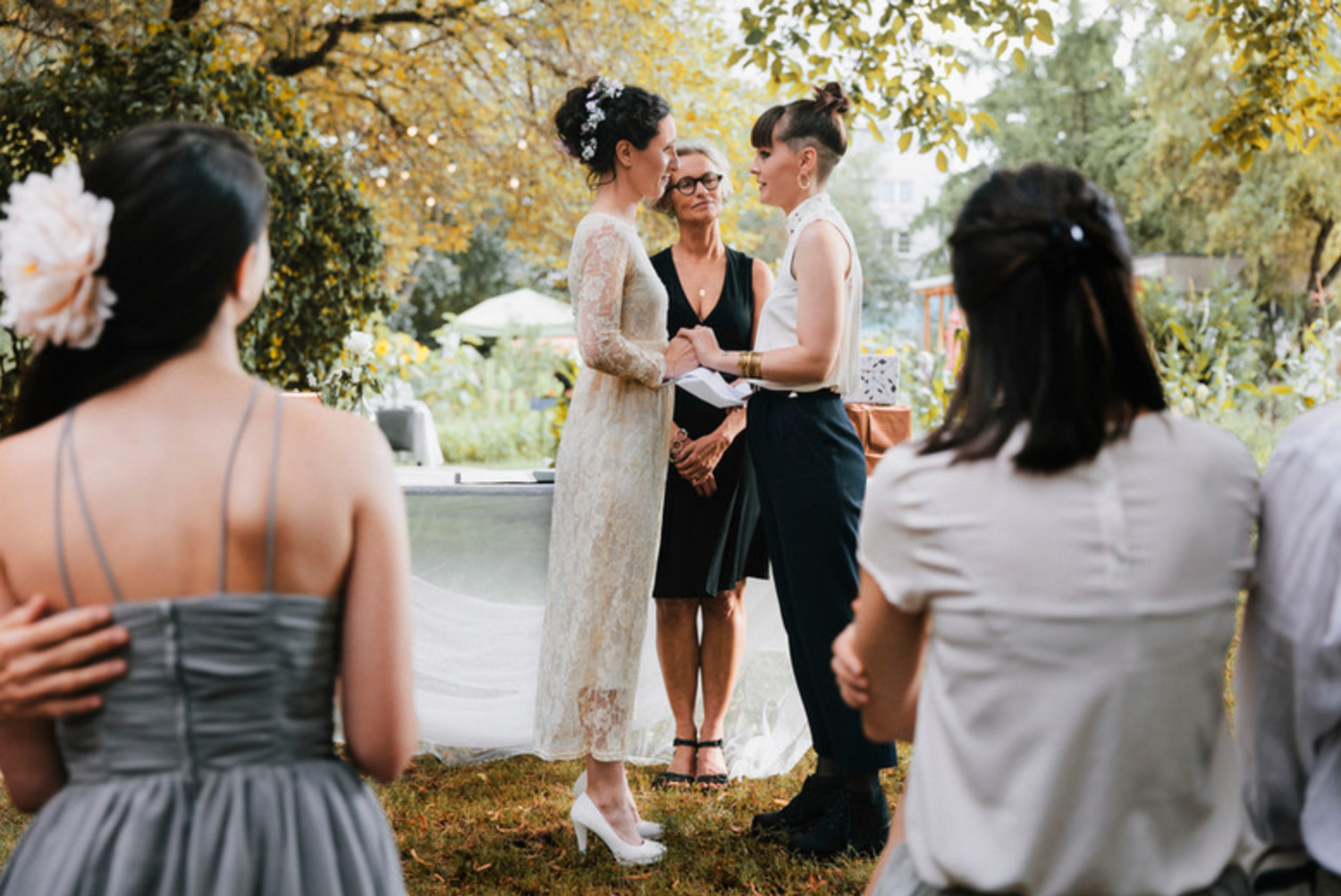 An intimate outdoor wedding ceremony under autumn trees with guests in gray dresses looking on.