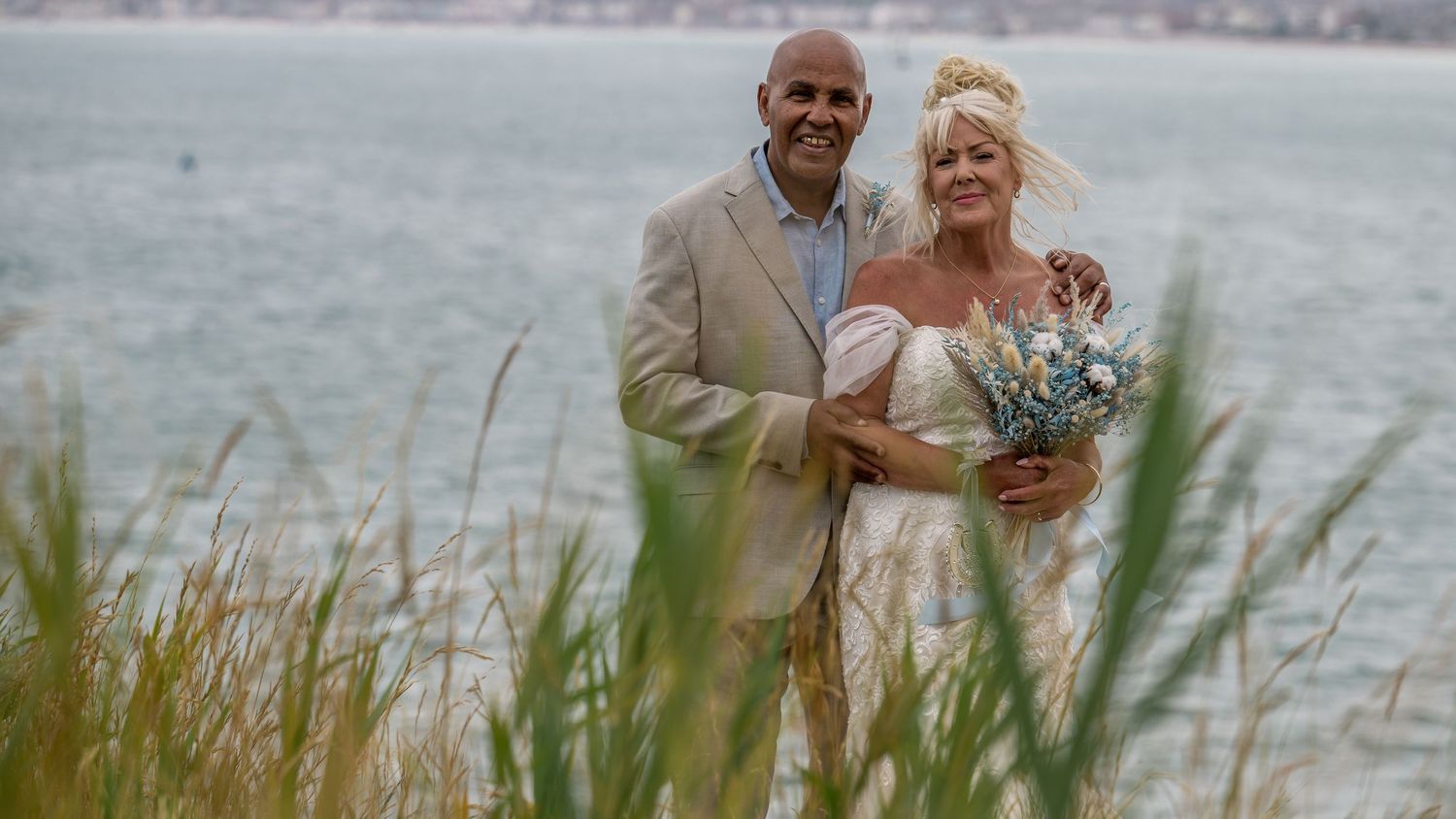 Newlyweds pose together by the ocean through tall grass on their wedding day.