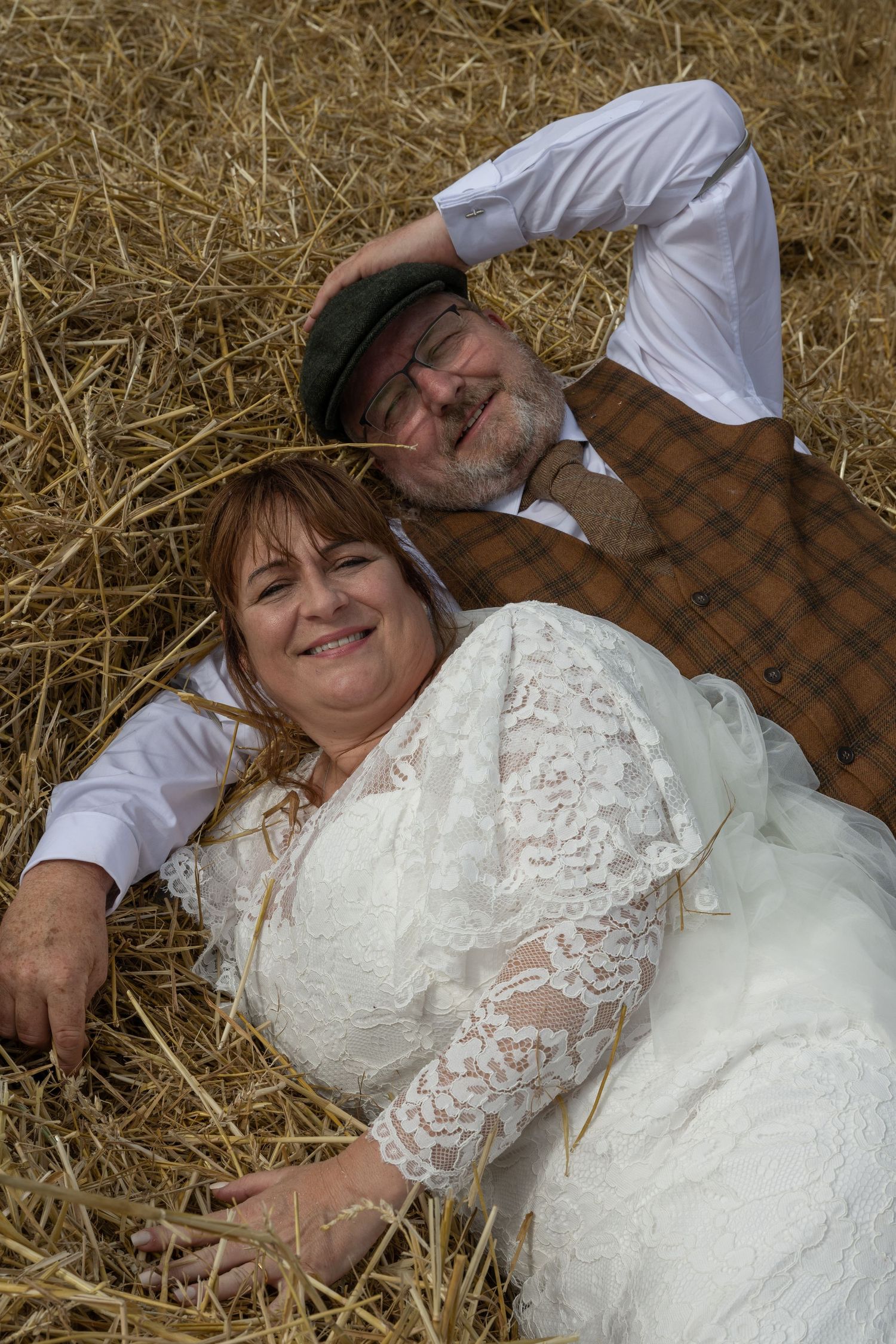 A couple in wedding attire lying together on hay bales in a rustic setting.