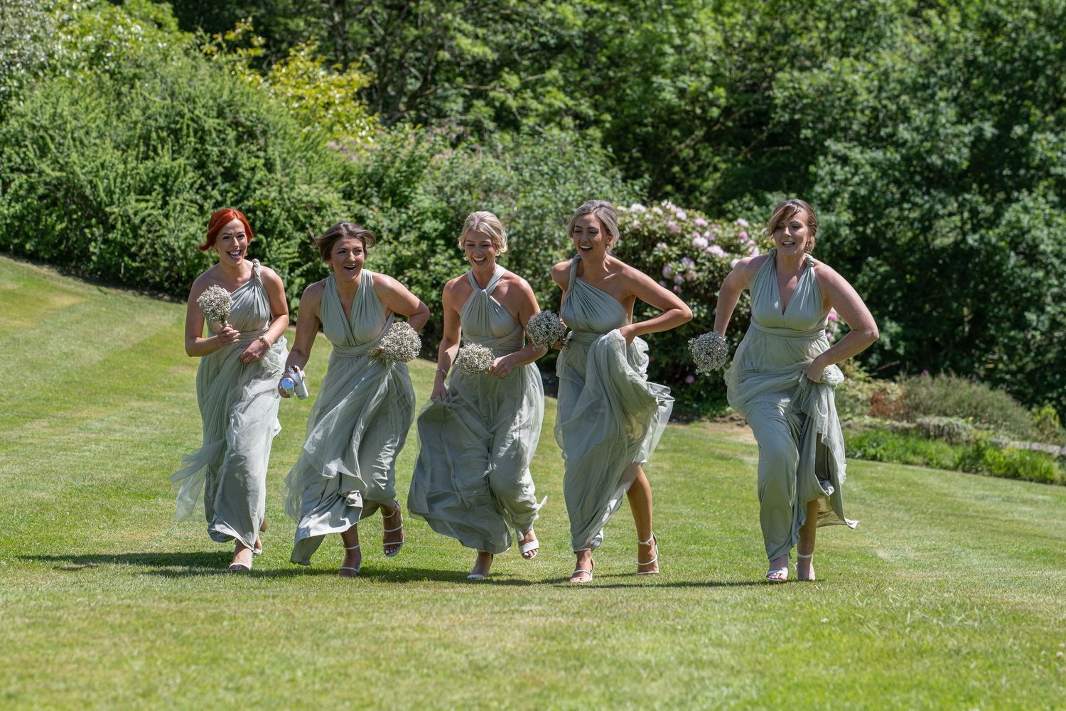 Bridesmaids in gray dresses running and laughing together on a sunny lawn.